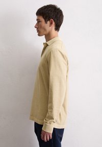 Młody mężczyzna stojący w profilu, ubrany w beżowy polo z długim rękawem i ciemne dżinsy, na tle gładkiej białej ściany.