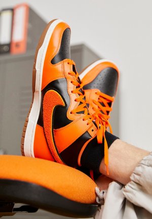 Baskets orange vif et noires avec des lacets orange portées par une personne reposant ses pieds sur une chaise orange dans un environnement intérieur.