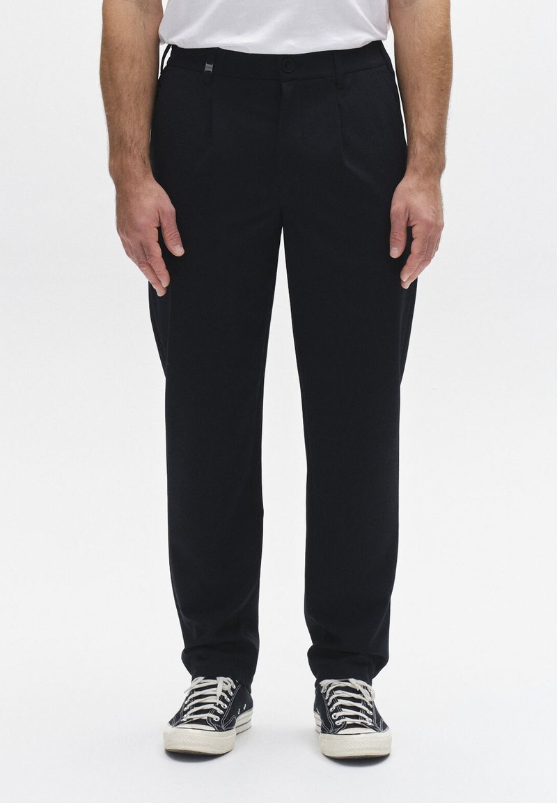 Férfi fekete egyenes szárú nadrágban és fekete, fehér fűzőjű tornacipőben, sima fehér háttér előtt állva.