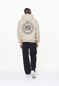 Sudadera beige con capucha y un logo negro redondo en la parte posterior, combinada con pantalones negros y zapatillas blancas. La tela parece suave y texturizada.