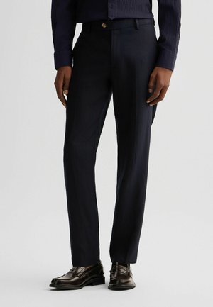 Pantaloni da completo blu navy con una texture liscia, design a gamba dritta, piega sottile e un bottone dorato in vita, abbinati a scarpe nere.