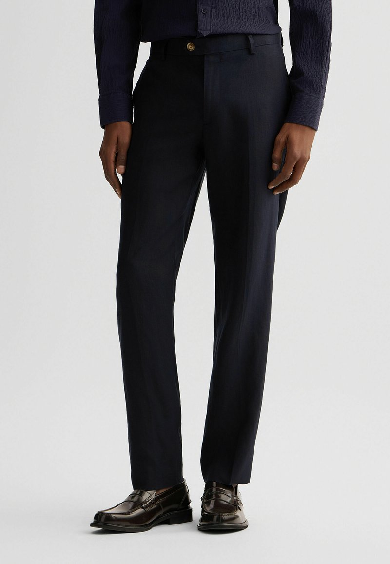 Pantaloni de costum navy cu o textură netedă, design cu picior drept, pliuri subtile și un nasture auriu la talie, asortați cu pantofi negri.