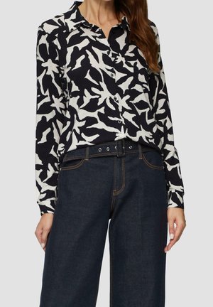 Femme portant une chemise à boutons imprimée en noir et blanc, rentrée dans un jean bleu foncé avec une ceinture noire.