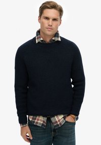 Granatowy sweter z prążkowanej dzianiny z okrągłym dekoltem, noszony na wierzchu koszuli flanelowej w kratę. Posiada rękawy raglanowe i teksturowany materiał.