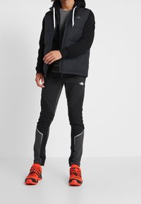 Zwarte gewatteerde vest over een grijze langemouwen shirt, gecombineerd met zwarte broek met grijze accenten en feloranje schoenen.