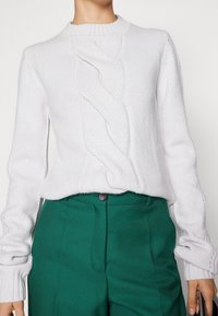 Bílý pletený svetr s texturovaným kabelkovým vzorem vpředu, s ribbovaným kulatým výstřihem a dlouhými rukávy, kombinovaný se zelenými kalhotami.