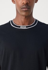 Juodas medvilninis marškinėlis su kontrastinga balta raukšlėta apykakle, ant kurios yra „BOSS“ logotipas baltos spalvos. Lygus paviršius ir prigludusi forma.