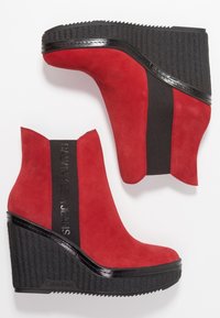 Czerwone botki na koturnie zamszowe z czarnymi elastycznymi panelami bocznymi i czarną teksturowaną platformą. Po boku znajduje się logo "Calvin Klein Jeans".