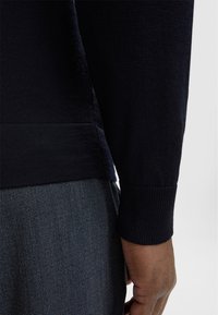 Marineblauer Pullover mit gerippten Bündchen und Saum, strukturiertem Stoff, Seitenschlitz, kombiniert mit dunkel gemusterten Hosen. Nahaufnahme des Ärmels und der Hand.