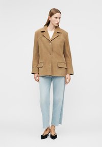 Vrouw die een tan suède blazer met twee knopen draagt, lichtblauwe jeans met afgewerkte zoom, en zwarte, puntige slingback schoenen, staand tegen een effen achtergrond.