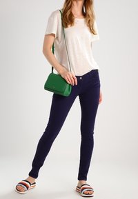 White, short-sleeved, lightweight top; navy slim-fit pants; green crossbody bag; striped slide sandals with black, white, and orange accents.