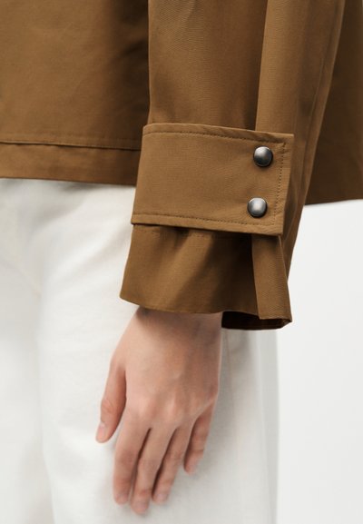 Μανίκι καφέ πουκάμισου με δύο μαύρα κουμπιά κοντά στην μανσέτα, φορεμένο από άτομο με ανοιχτόχρωμο δέρμα, με το χέρι να ακουμπάει δίπλα σε λευκό παντελόνι.