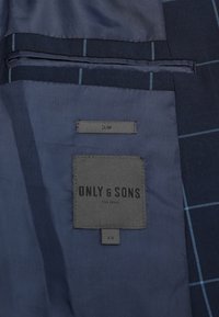 Wnętrze granatowej marynarki z teksturowanej tkaniny. Zawiera etykietę pielęgnacyjną oraz metkę z logo "ONLY & SONS", rozmiar 48, w niebieską kratkę.