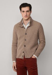 Béžový pletený cardigan s golierom, s čiernymi gombíkmi a textúrovanou väzbou, v kombinácii s károvanou košeľou a hnedými nohavicami.