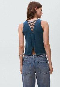 Teal stickad linne med en snörad design på baksidan. Har en kortad längd och rå fåll, kombinerat med högmidjade blå denimjeans.
