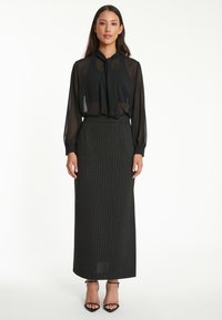 Zwarte doorschijnende blouse met een nekstrik, gecombineerd met een lange zwarte rok met streepjesmotief. Model draagt zwarte gekruiste hakken.