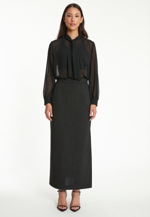 Schwarze durchsichtige Bluse mit Halsknoten, kombiniert mit einem langen schwarzen Rock mit Nadelstreifen. Das Model trägt schwarze Riemchensandalen.