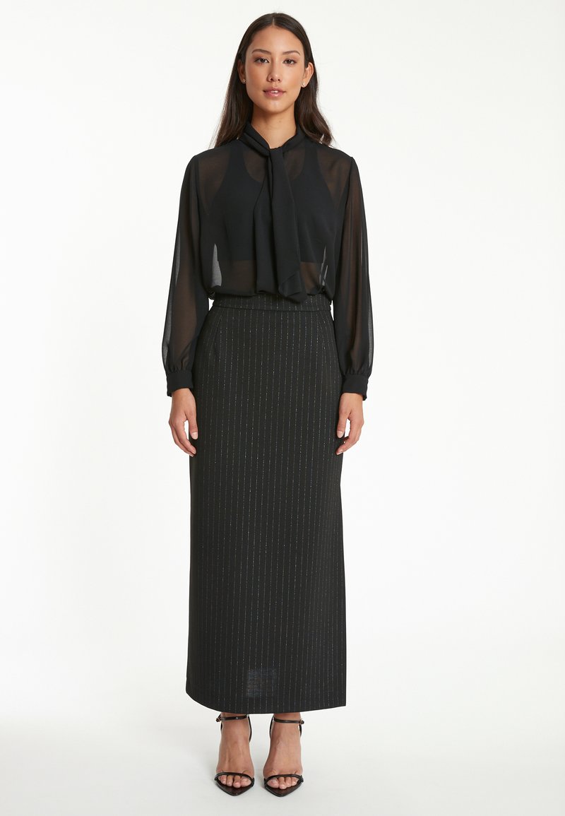 Zwarte doorschijnende blouse met een nekstrik, gecombineerd met een lange zwarte rok met streepjesmotief. Model draagt zwarte gekruiste hakken.