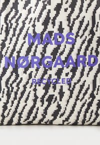 Perdirbtos medžiagos krepšys su juodai baltu zigzago raštu, ant kurio stora violetine spalva užrašyta "MADS NØRGAARD" ir "RECYCLED".