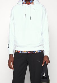 White hoodie with drawstrings, front pocket, and black logo detail; paired with black pants featuring a colorful patterned waistband.