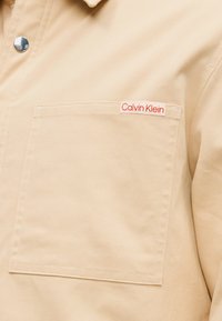 Μπεζ βαμβακερό πουκάμισο με τσέπη στο στήθος και κόκκινη ετικέτα με το λογότυπο Calvin Klein. Λεία υφή με κλασικό γιακά και κούμπωμα με κουμπιά πατιλέτα.