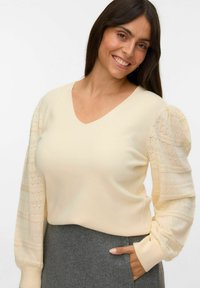 Cremfarbener Pullover mit V-Ausschnitt und strukturierten Spitzen-Puffärmeln. Der Körper ist glatt und wird mit einem grauen Rock kombiniert.