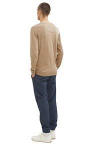 Uomo in piedi con le mani in tasca, che indossa un maglione beige, pantaloni jogging blu e scarpe da ginnastica bianche, visto di spalle su uno sfondo bianco.