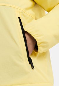 Giacca a vento gialla con una tasca con cerniera nera, texture liscia, polsini elasticizzati e design minimalista.