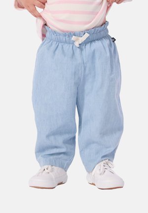 Kleinkind trägt lockere hellblaue Hose mit gerafftem Bund und weißen Turnschuhen, hebt rosa gestreiftes Oberteil hoch.