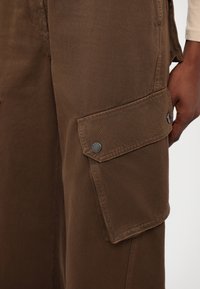Pantalon cargo marron avec une poche latérale à fermeture par bouton, fabriqué en tissu texturé avec des détails de coutures.