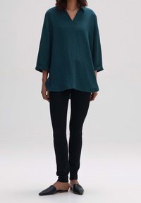 Turkusowa strukturalna bluzka z długim rękawem i dekoltem w kształcie litery V, w zestawie z czarnymi slim fit spodniami i płaskimi czarnymi butami, na neutralnym tle.