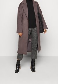 Lång, grå kappa med öppen front, gjord av ull, med sänkt axelparti. Kombination med svart polotröja, rutiga byxor och knähöga svarta stövlar.