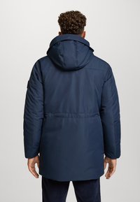 Uomo con capelli ricci che indossa un cappotto invernale blu scuro con cappuccio, in piedi con le mani rilassate lungo i fianchi, visto da dietro.
