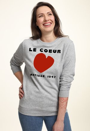 Kvinde smiler og kigger op iført en grå sweatshirt med et rødt hjerte og teksten "LE COEUR MATISSE, 1947" samt blå jeans.