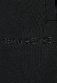 Fekete szövet, amely texturált kötésű és hímzett szöveget tartalmaz: "9N1M SENSE." Finom részletekkel és látható minták vagy hardver nélkül.