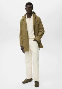 Vaaleanoliivinvihreä kevyt takki, kerma-valkoinen polo, valkoiset housut ja ruskeat mokkakengät. Takissa on huppu ja etutaskut.