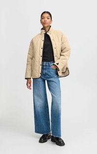 Veste matelassée beige avec un col haut, des accents noirs et des boutons-pression, associée à un jean en denim bleu à jambes larges et des chaussures noires.