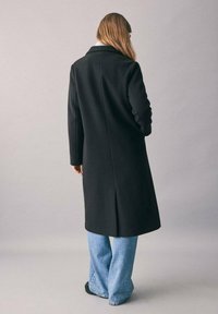 Lungo cappotto nero realizzato in un materiale morbido, con colletto a revers, vestibilità strutturata e spacco posteriore. Indossato sopra jeans larghi blu.
