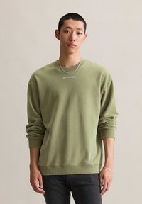 Jonge man in een licht olijfgroen sweatshirt met de tekst "MARC O'POLO DENIM", zwarte broek, zilveren ketting en armband, staande.
