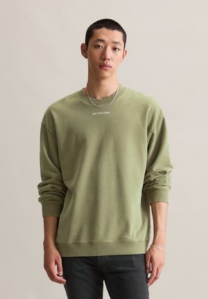 Jeune homme portant un sweat-shirt vert olive clair avec le texte "MARC O'POLO DENIM", un pantalon noir, un collier en chaîne argenté et un bracelet, se tenant debout.
