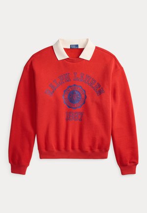 Femme avec de longues tresses portant un sweat-shirt rouge Ralph Lauren et un pantalon large bleu marine, assise sur un tabouret en bois devant un fond uni.