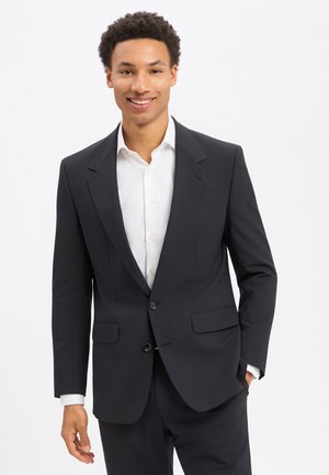 Lächelnder Mann in einem schwarzen Anzug und weißem Hemd steht mit einer Hand in der Tasche vor einem schlichten weißen Hintergrund.