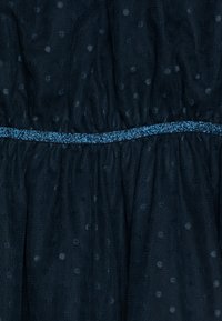Tengerészes ruha anyag pöttyös textúrával és ráncolt derékkal. Jellemzője egy kék csillogó sáv, amely kiemeli a derékvonalat.