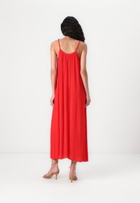 Robe maxi rouge, avec des bretelles spaghetti et un dos froncé. Texture lisse, design fluide et ourlet droit. Portée avec des sandales à talons couleur sable.