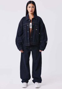Set composto da giacca e pantaloni in denim scuro, caratterizzato da una vestibilità ampia, cuciture a contrasto, tasche sul petto e leggeri dettagli con il logo. Sneakers bianche completano il look.