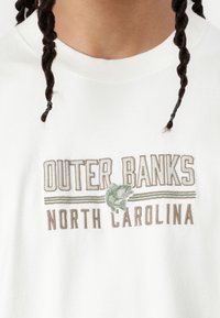 Fehér rövidujjú póló, amelyen barna és zöld hímzett szöveggel szerepel az "OUTER BANKS" és a "NORTH CAROLINA", hal mintával és csíkokkal.
