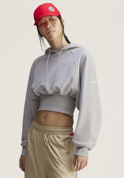 Joven mujer con gorra roja, sudadera gris recortada y pantalones deportivos beige, de pie frente a un fondo liso y claro.