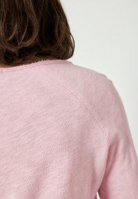 Hnědé vlasy po ramena přes záda světle růžového trička s dlouhým rukávem a ohrnutým výstřihem na jednobarevném pozadí.