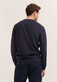 Ein Mann trägt einen dunkelblauen Langarmpullover und eine dunkel gemusterte Hose und steht mit dem Rücken zu einem schlichten hellen Hintergrund.