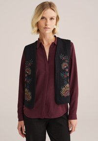 Zwarte gewatteerde gilet met bloemenborduurwerk in rood, geel en groen, gedragen over een bordeaux shirt. Heeft een recht model en een gedetailleerd patroon.